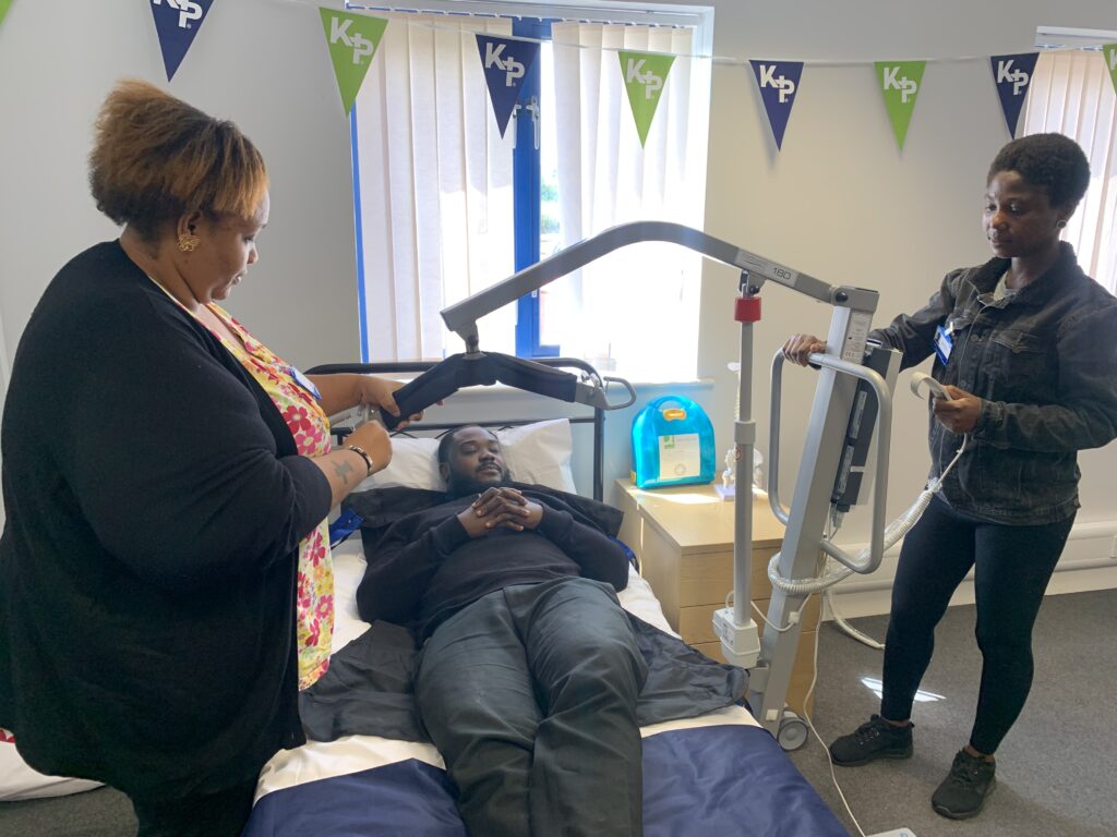 Two female care staff practice using a hoist to move a patient from the bed to a chair.