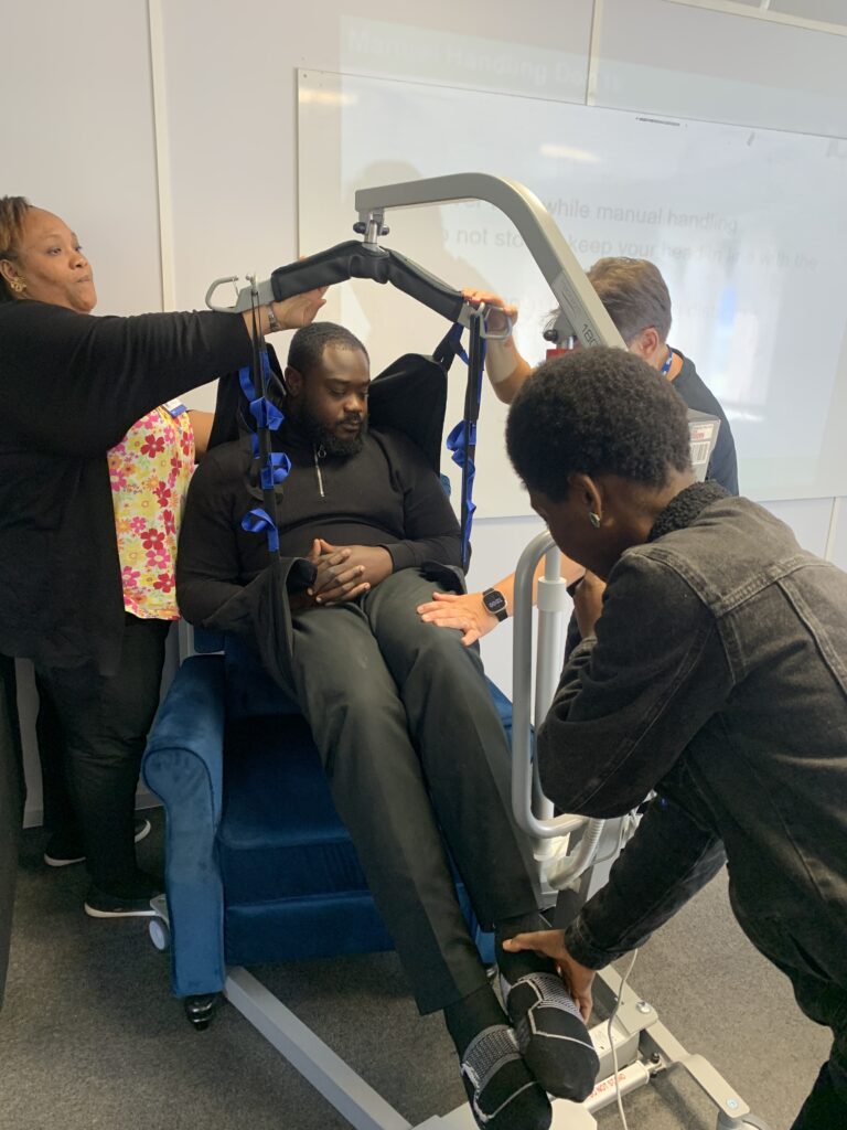 A patient is transferred to chair using a hoist, practiced by two Kare Plus carers under the supervision of their trainer.